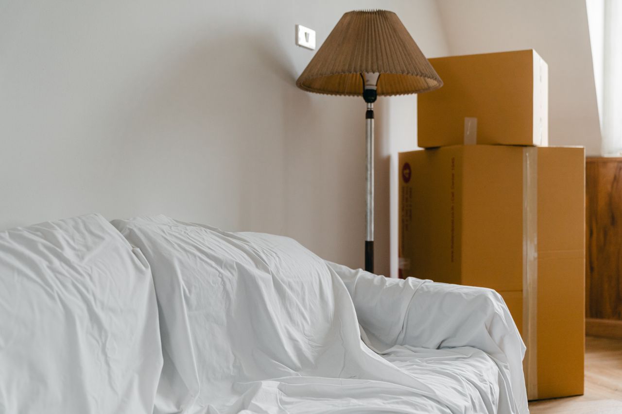 creative-02 Covered with sheet sofa and floor lamp and packed cardboard boxes in light spacious room of new contemporary house