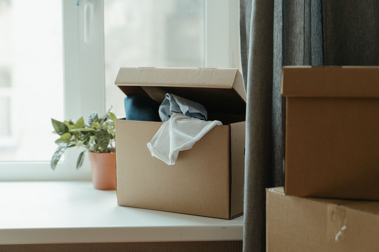 Services-02 Open cardboard box with clothes by window, symbolizing moving or relocation.